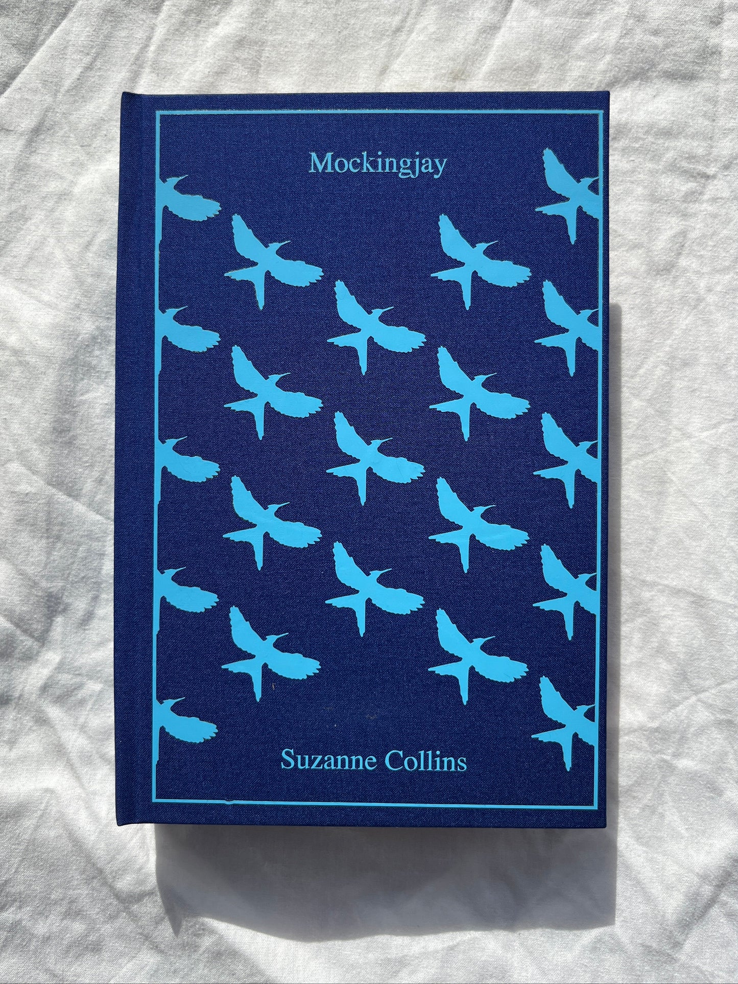 The book cover of 'Mockingjay' by Suzanne Collins, featuring a dark navy blue background with a repeating pattern of light blue swallow birds, displayed on a wrinkled white fabric surface.