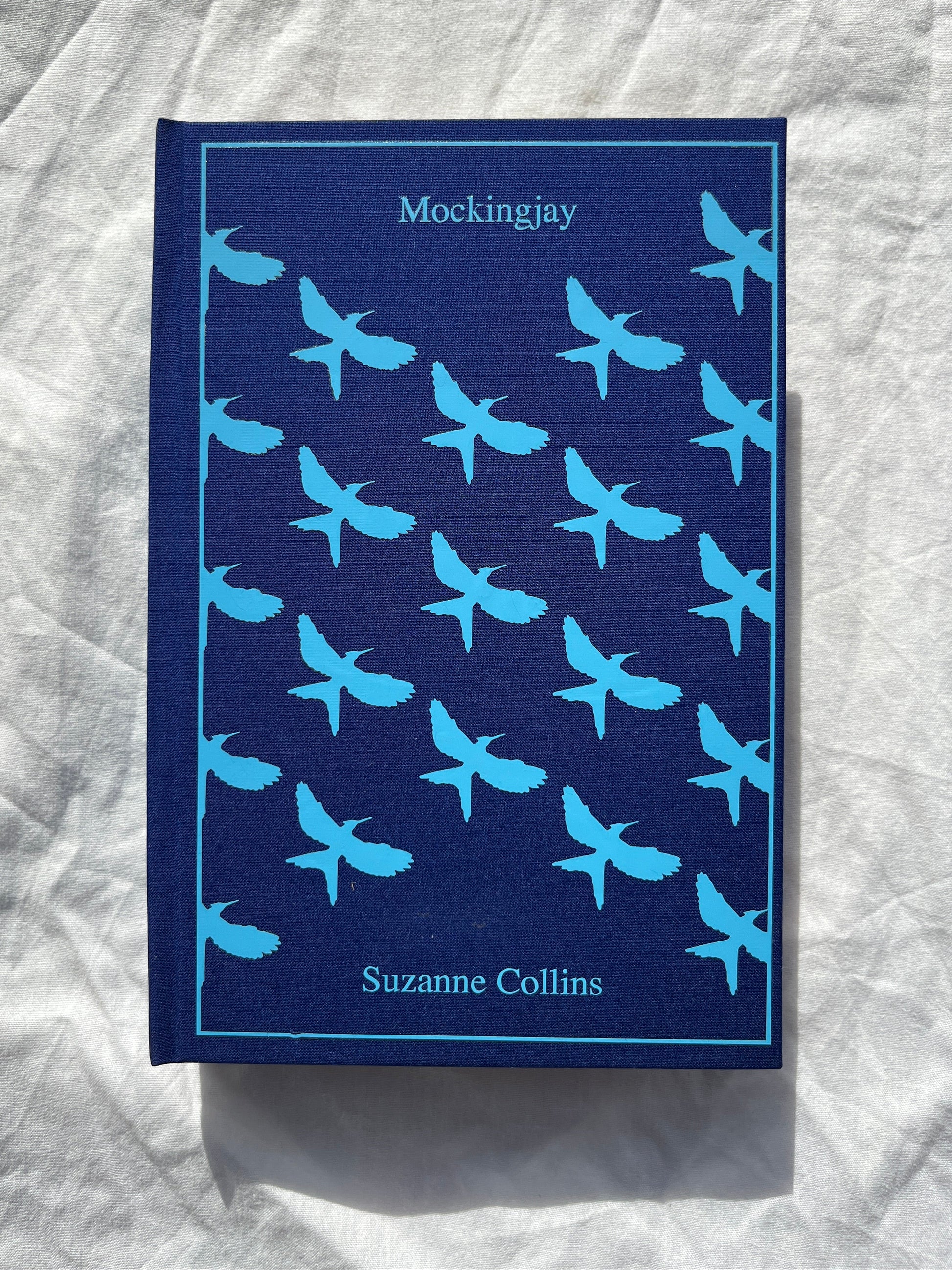 The book cover of 'Mockingjay' by Suzanne Collins, featuring a dark navy blue background with a repeating pattern of light blue swallow birds, displayed on a wrinkled white fabric surface.