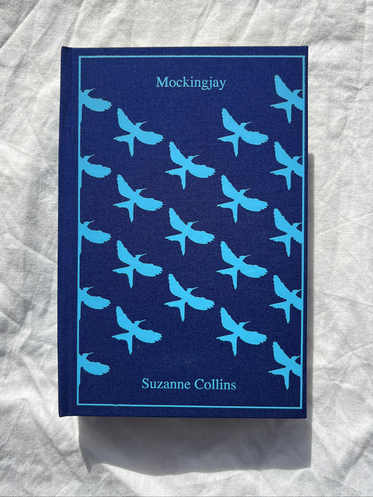 The book cover of 'Mockingjay' by Suzanne Collins, featuring a dark navy blue background with a repeating pattern of light blue swallow birds, displayed on a wrinkled white fabric surface.