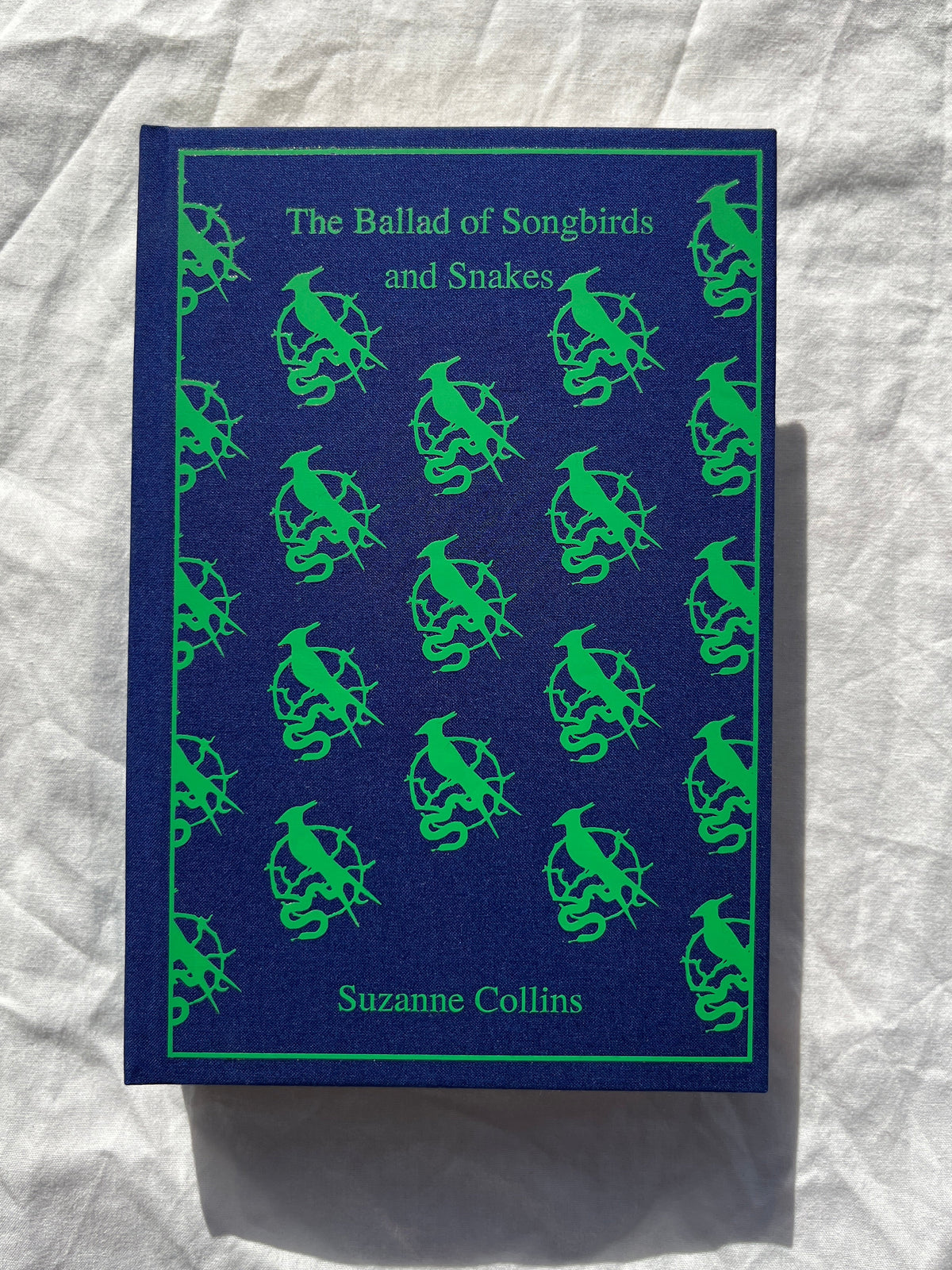 The Ballad of Songbirds and Snakes book by Suzanne Collins, featuring a dark blue cover with a repeating green pattern of birds and snakes, photographed on a wrinkled white fabric background.