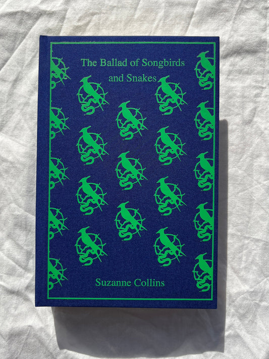 The Ballad of Songbirds and Snakes book by Suzanne Collins, featuring a dark blue cover with a repeating green pattern of birds and snakes, photographed on a wrinkled white fabric background.